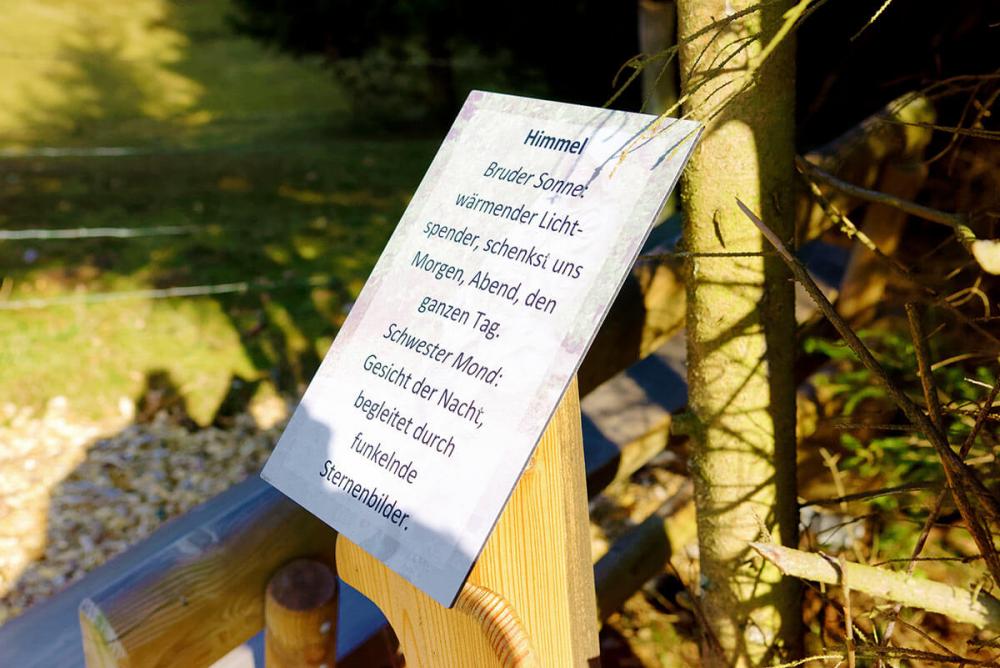 Hinweisschild mit einem Gedicht namens „Himmel“ auf dem Assisi-Pfad. Text: „Bruder Sonne: wärmender Lichtspender, schenkst uns Morgen, Abend, den ganzen Tag. Schwester Mond: Gesicht der Nacht, begleitest durch funkelnde Sternenbilder.“