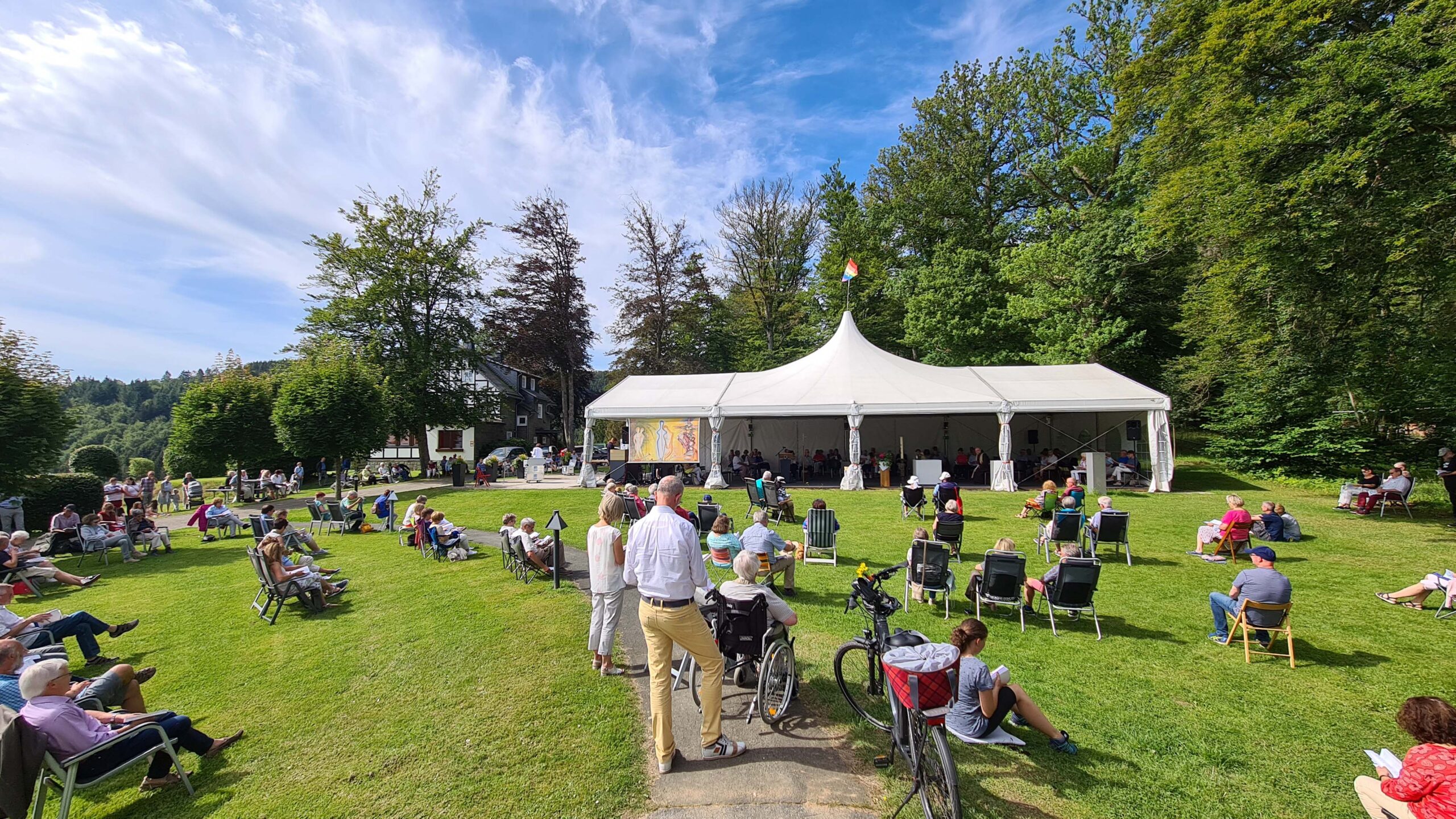 Gottesdienst im Freien vor einer Zeltkirche, Besucher sitzen mit Abstand auf einer Wiese inmitten grüner Natur.