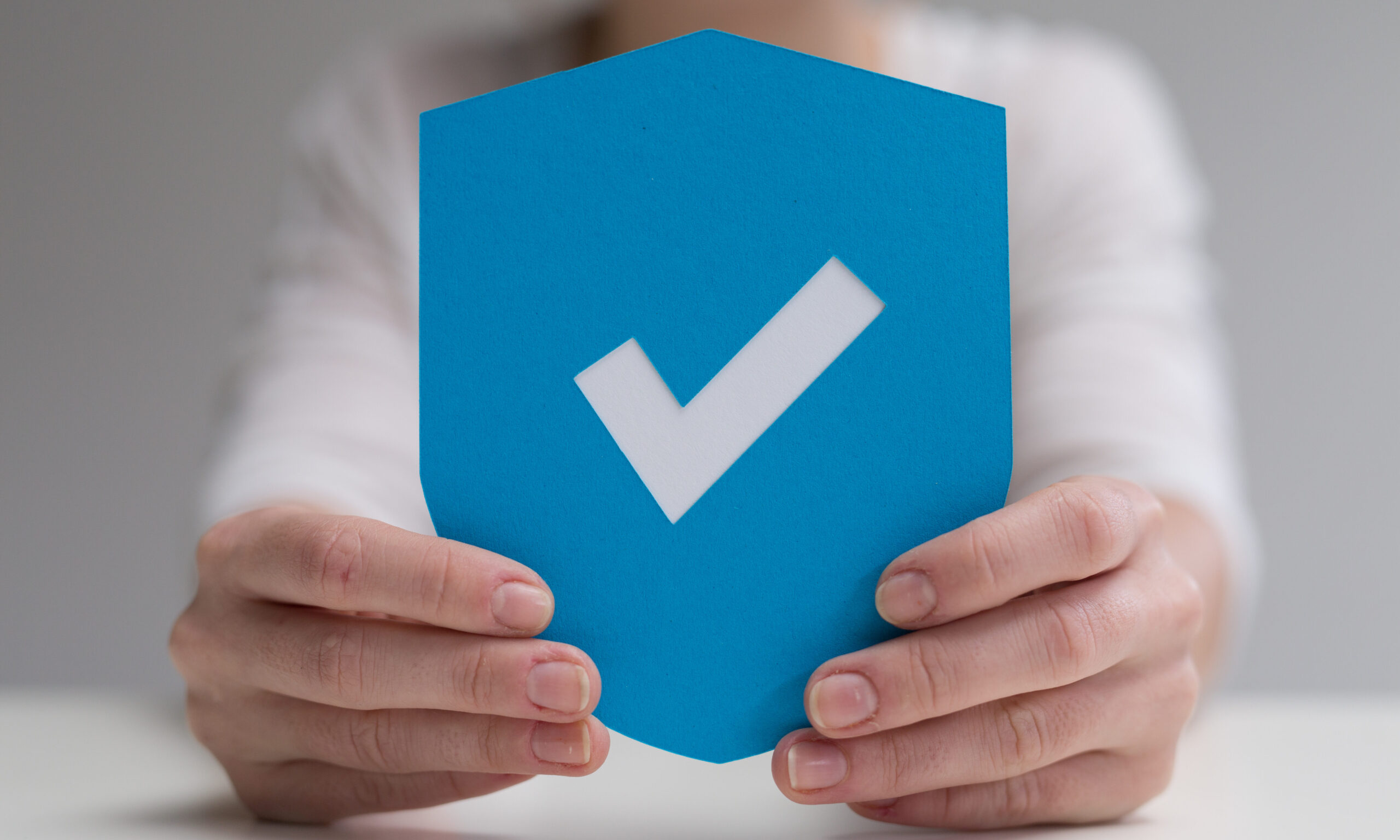 Hände halten ein blaues Schild mit einem weißen Häkchen als Symbol für Sicherheit, Schutz oder Bestätigung.