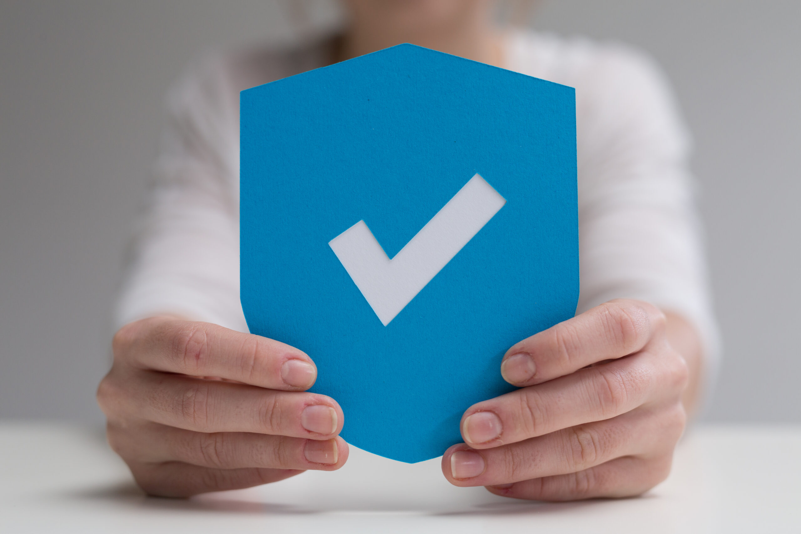 Hände halten ein blaues Schild mit einem weißen Häkchen als Symbol für Sicherheit, Schutz oder Bestätigung.