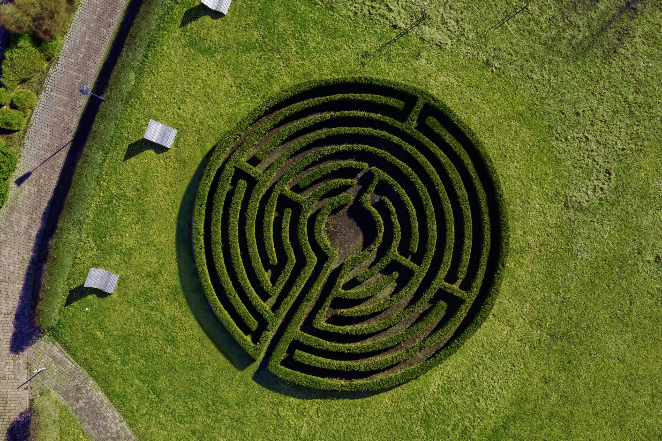 Draufsicht auf ein kreisförmig angelegtes Heckenlabyrinth inmitten einer grünen Wiese; umliegende Wege und einzelne Bänke laden zum Verweilen und Erkunden ein.