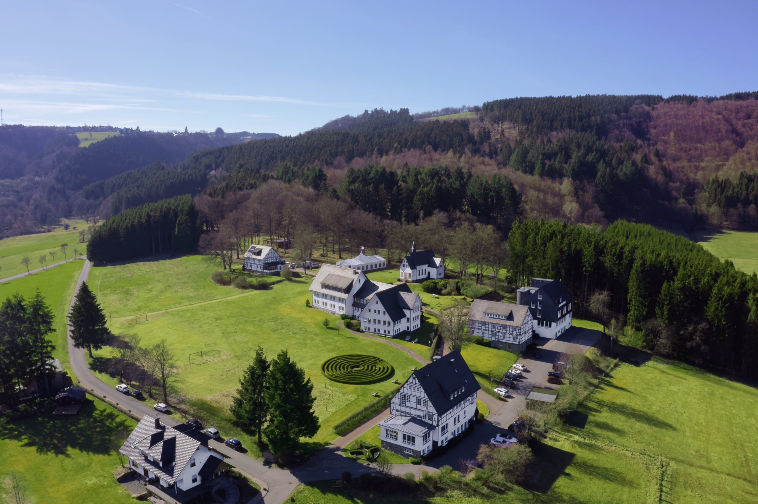 Luftaufnahme einer kleinen ländlichen Siedlung mit mehreren weiß gestrichenen Häusern, umgeben von grünen Wiesen und dicht bewaldeten Hügeln; im Vordergrund ein geometrisch angelegter Kreisgarten, daneben eine schmale Straße mit wenigen parkenden Autos unter klarem blauem Himmel.