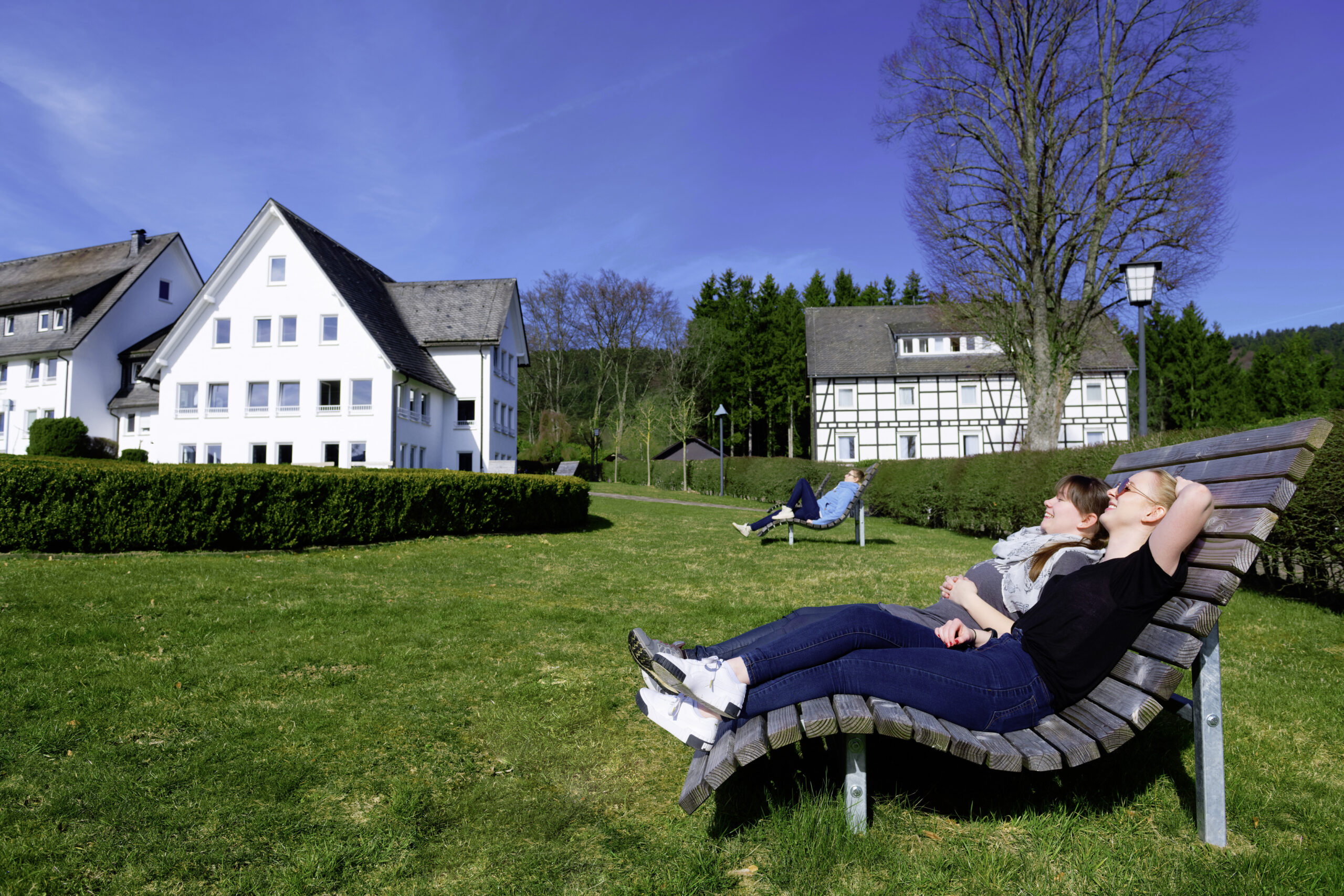 Zwei Frauen entspannen auf Holzliegen in einer gepflegten Grünanlage vor mehreren hellen Gebäuden; sie genießen die Sonne und blicken entspannt in den blauen Himmel, während im Hintergrund weitere Menschen und ein ruhiges, ländliches Umfeld mit Bäumen und Wiesen zu sehen sind.