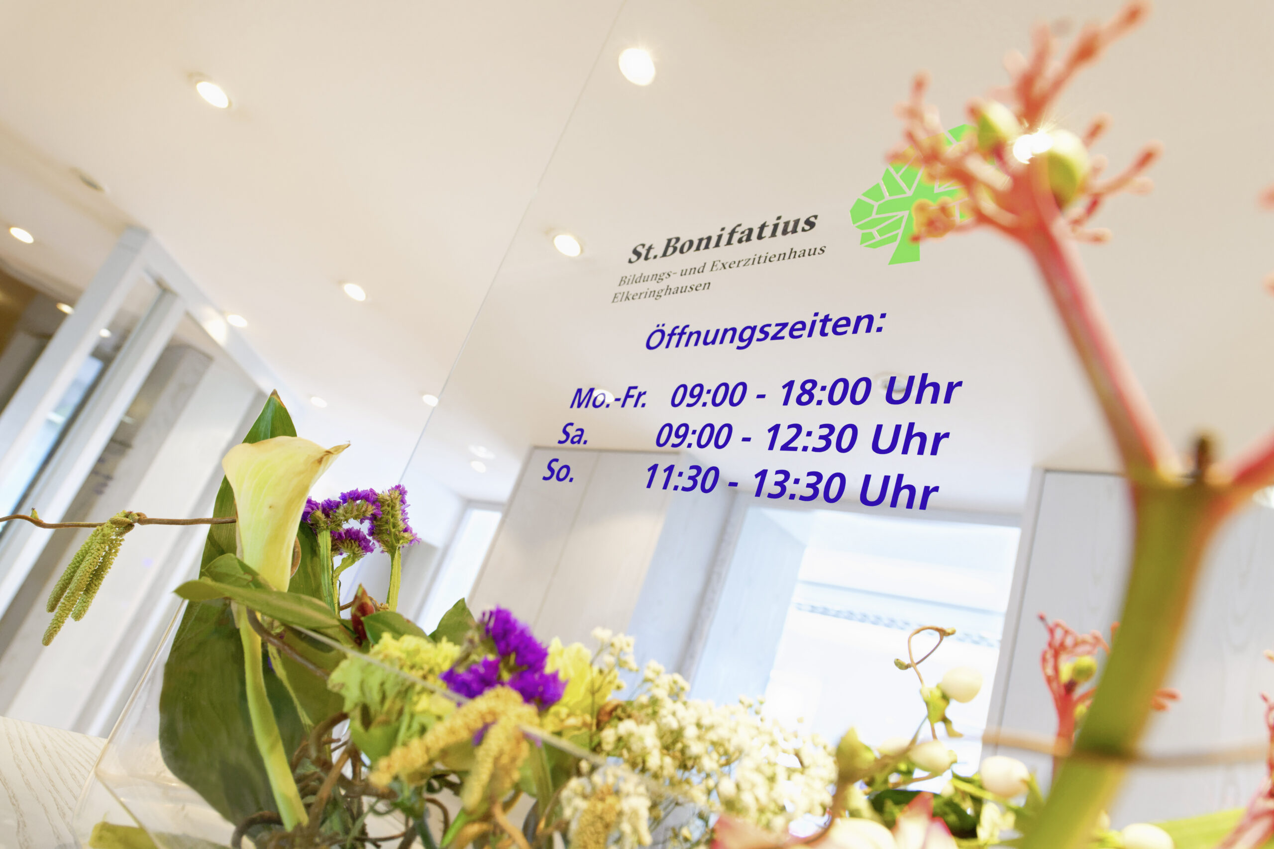 Innenansicht des Bildungs- und Exerzitienhauses St. Bonifatius mit Blumendekoration im Vordergrund und sichtbar angebrachten Öffnungszeiten an einer Glaswand.