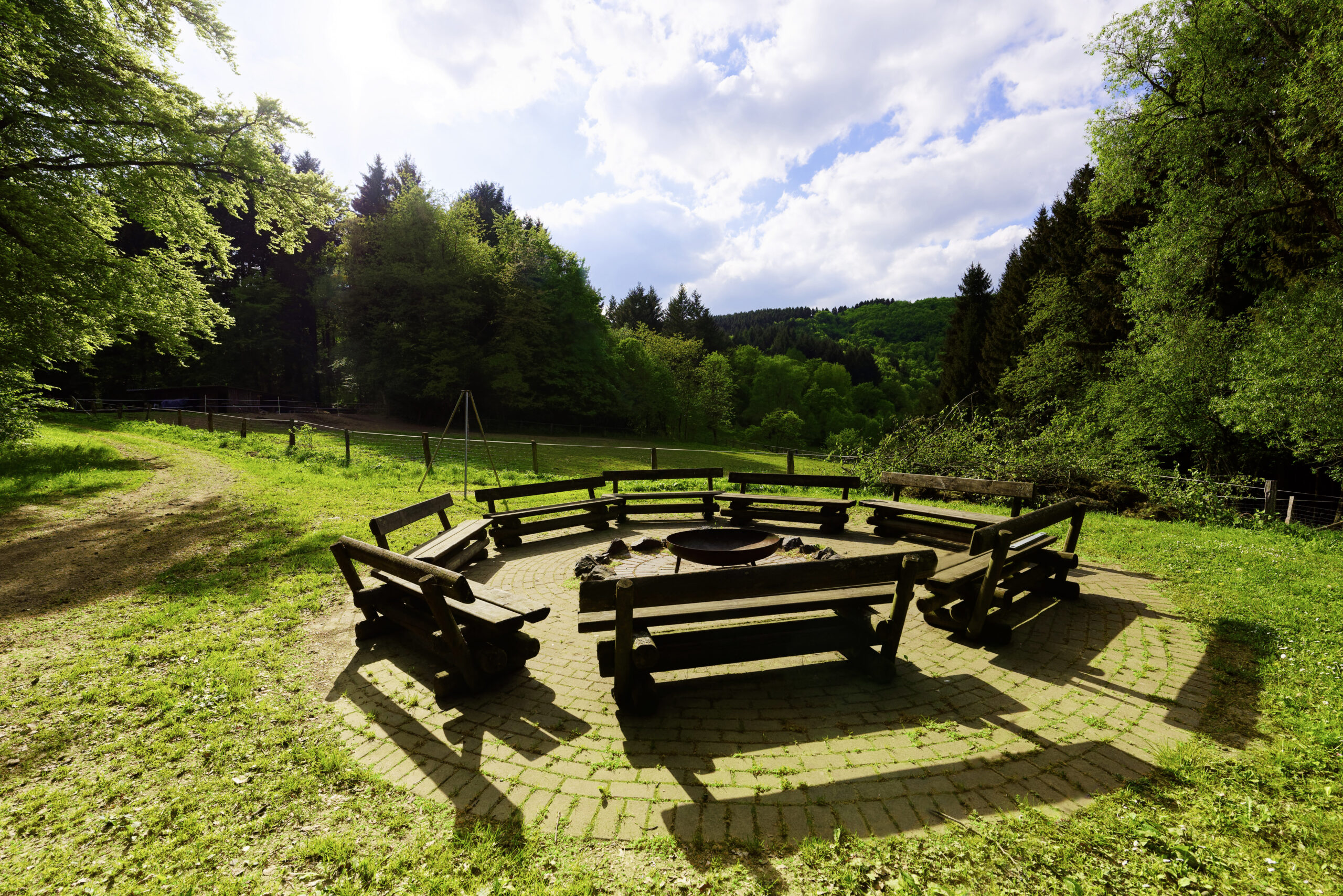 Runder Sitzplatz mit Holzbänken um eine Feuerstelle im Grünen, umgeben von Wiesen und Wald.