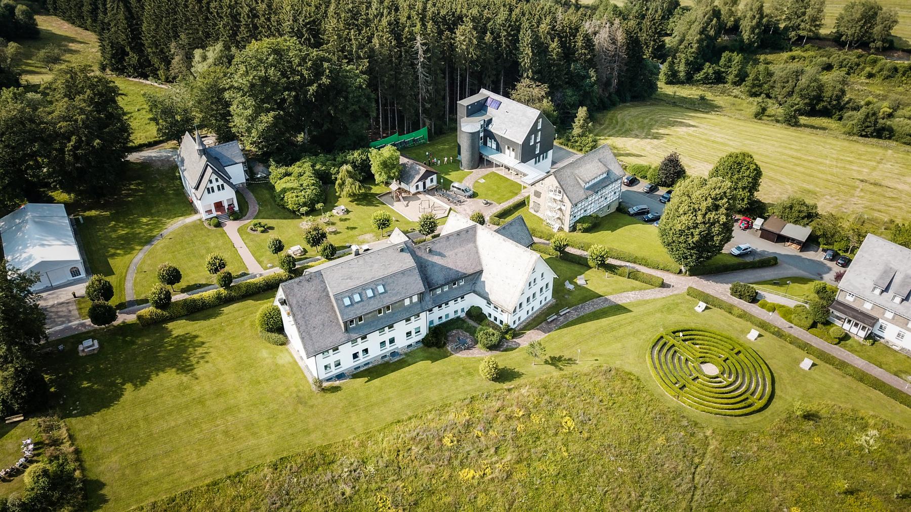 Luftaufnahme eines weitläufigen Gebäudekomplexes mit Parkanlagen, Labyrinth und umgebenden Wiesen am Waldrand.