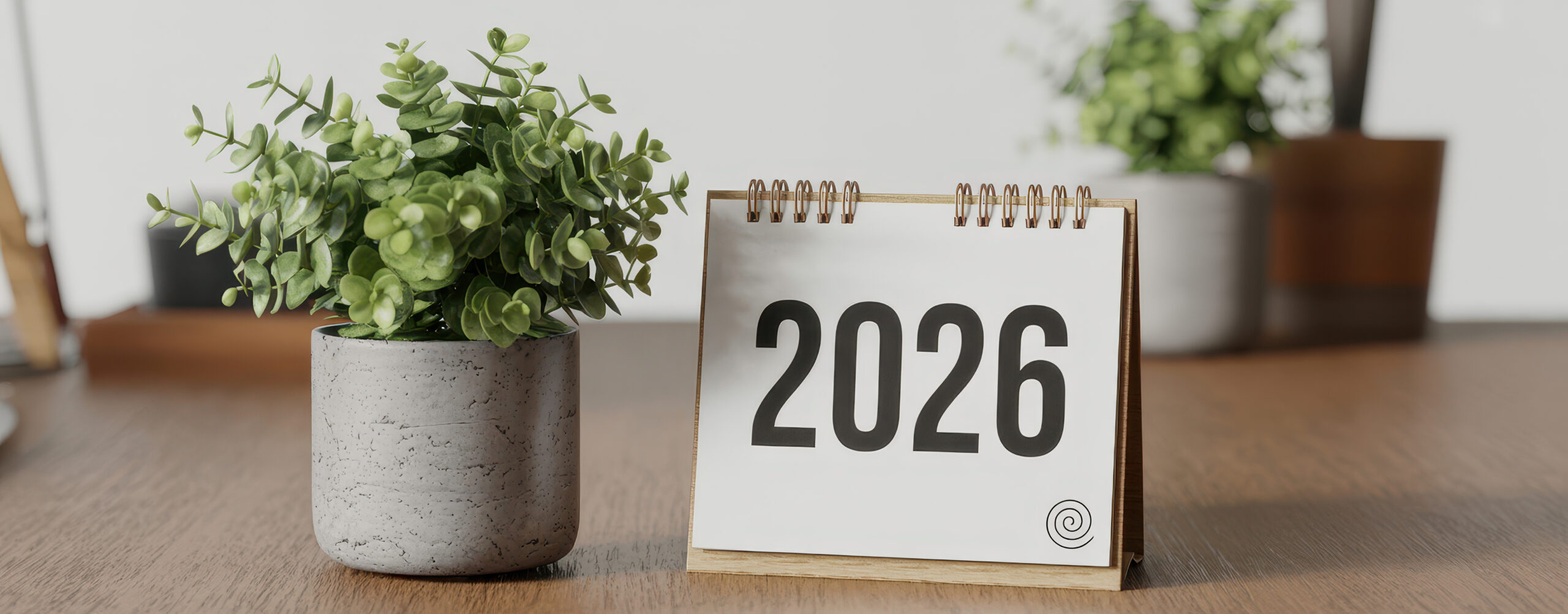 Tischkalender mit der Jahreszahl 2026 neben einer Topfpflanze auf einem Schreibtisch.