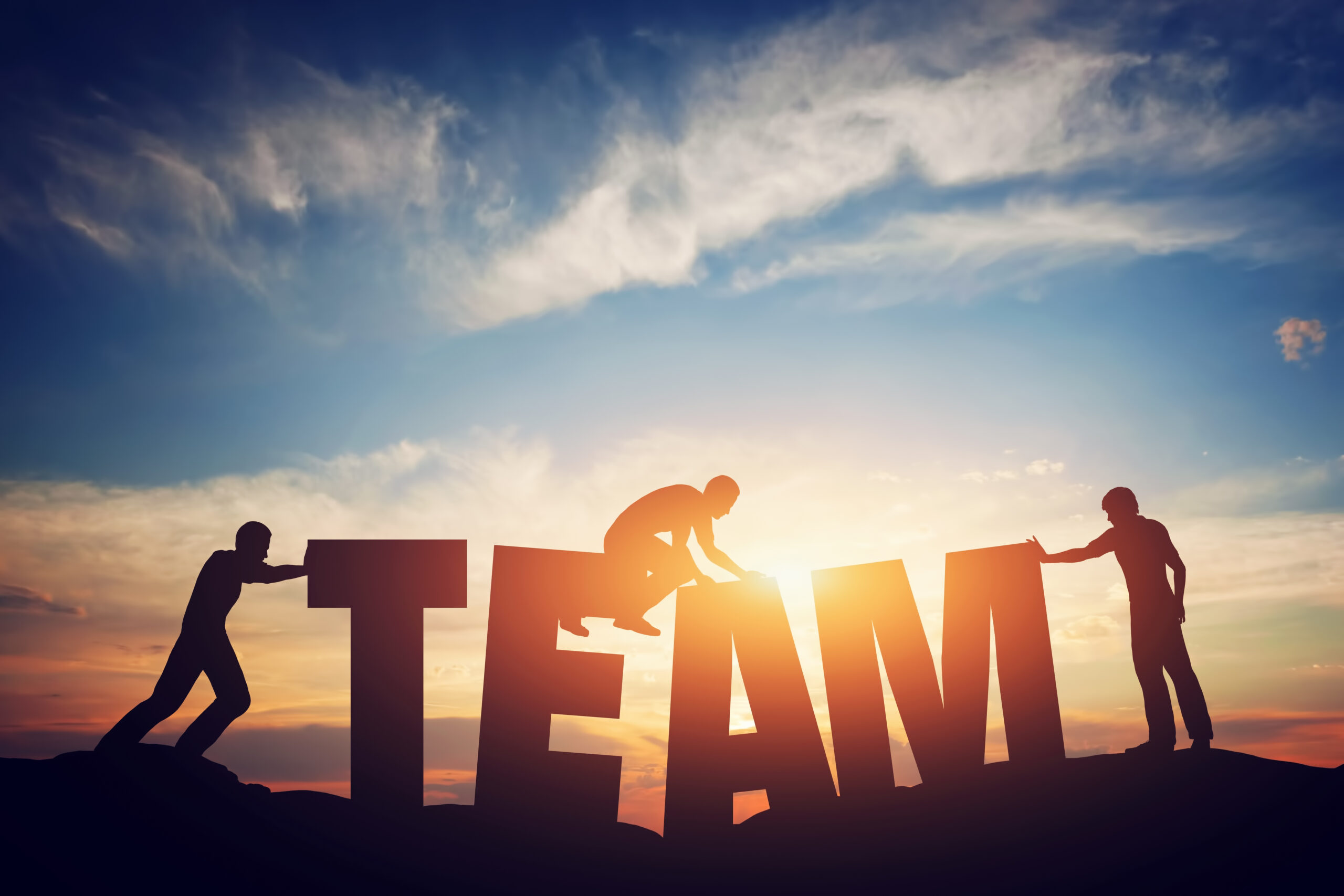 Silhouetten von drei Personen, die gemeinsam große Buchstaben zum Wort „TEAM“ aufrichten, vor einem Sonnenuntergang.