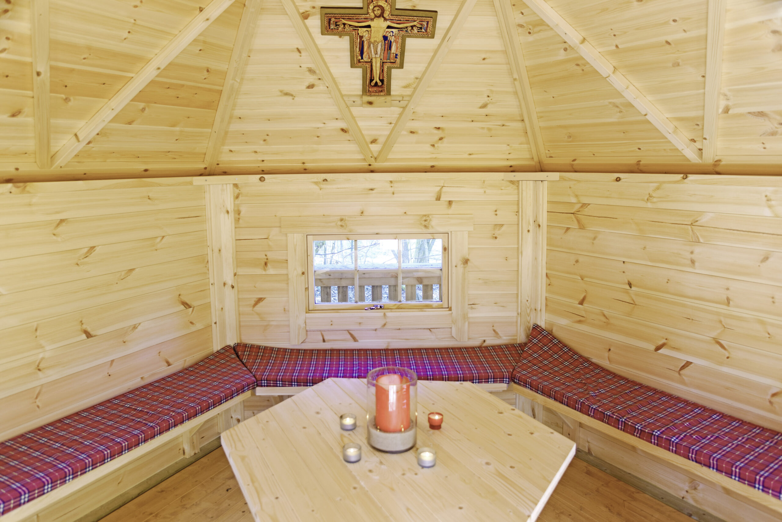 Innenraum einer hölzernen Gebets- oder Meditationshütte mit Sitzbänken, Tisch, Kerzen und Kreuz an der Wand.