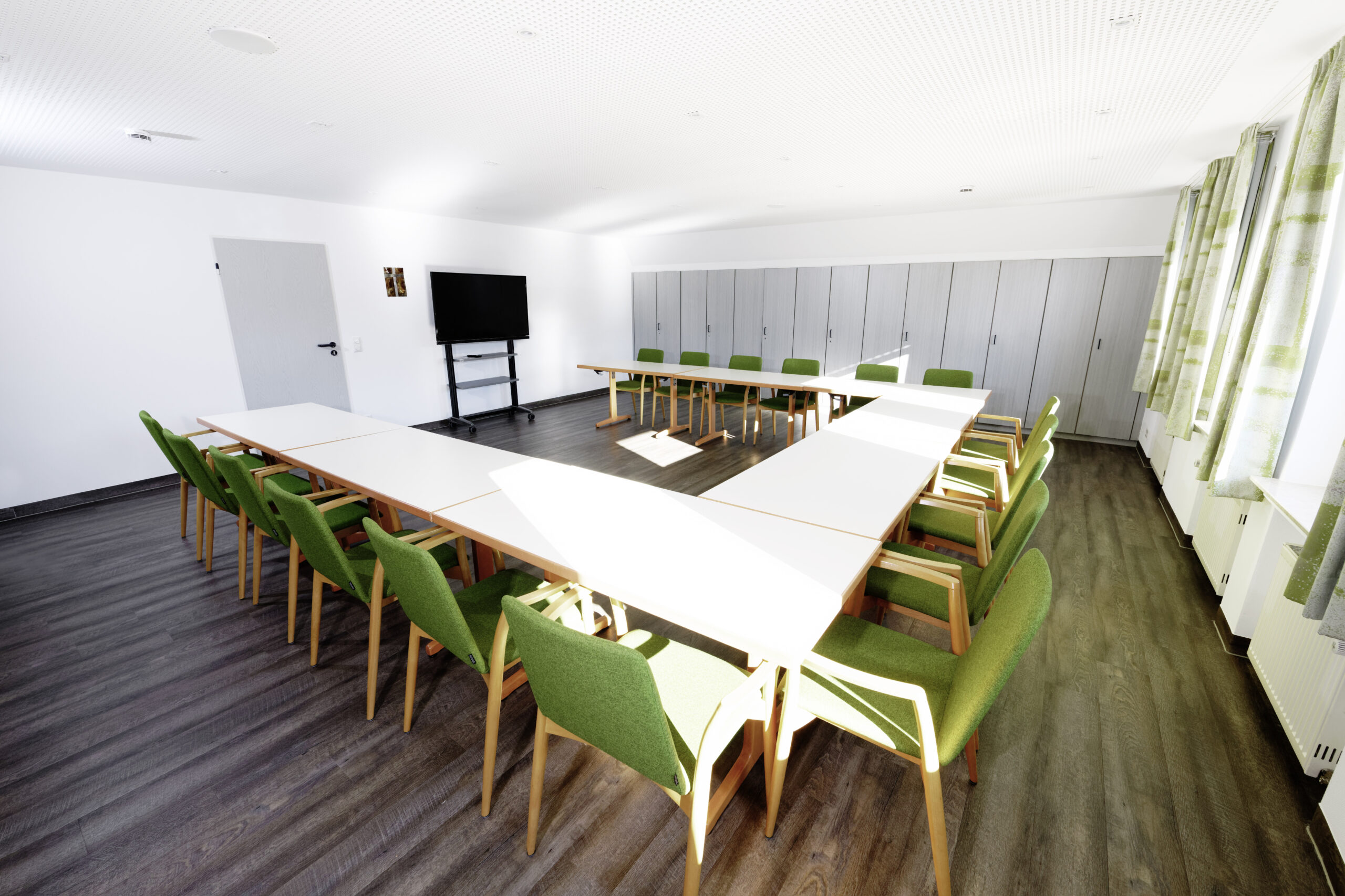 Heller Seminarraum mit U-förmig angeordneten Tischen, grünen Stühlen und Bildschirm für Besprechungen, Workshops und Schulungen.