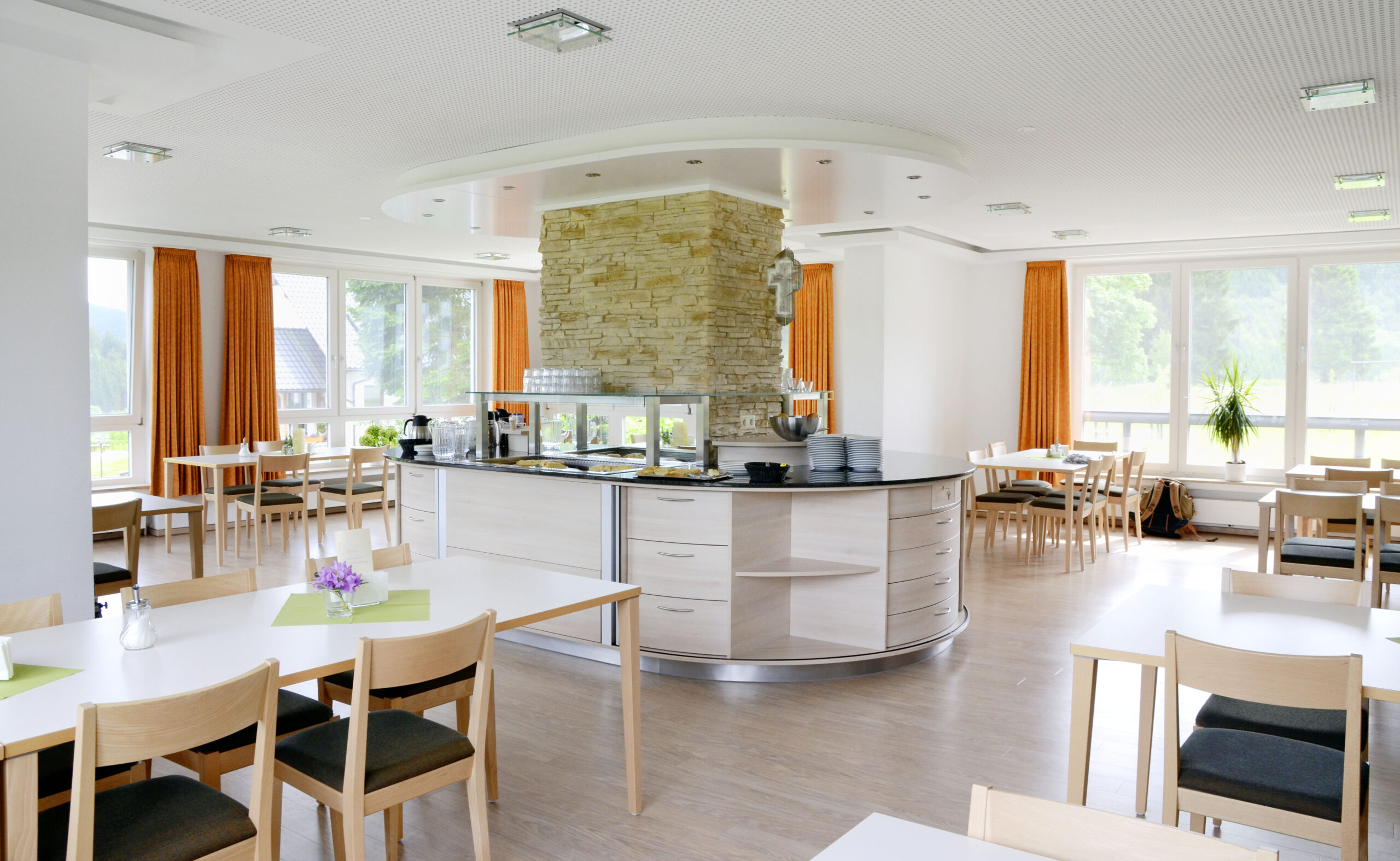 Heller Speisesaal mit zentralem Buffetbereich, Sitzgruppen und großen Fenstern mit Blick ins Grüne.