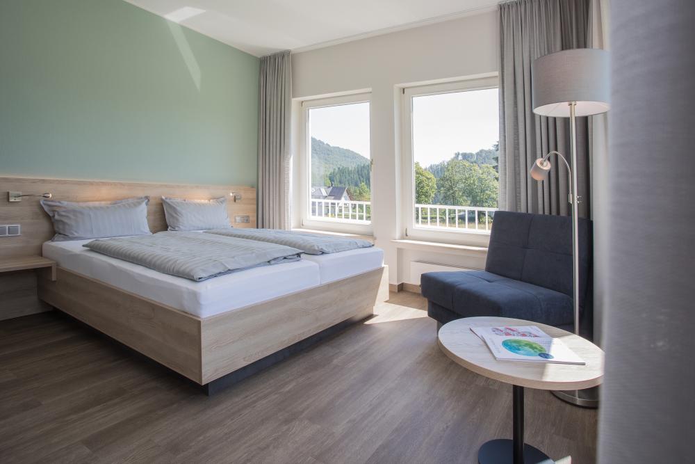Modern eingerichtetes Gästezimmer mit Doppelbett, Sitzgelegenheit und großen Fenstern mit Blick ins Grüne.