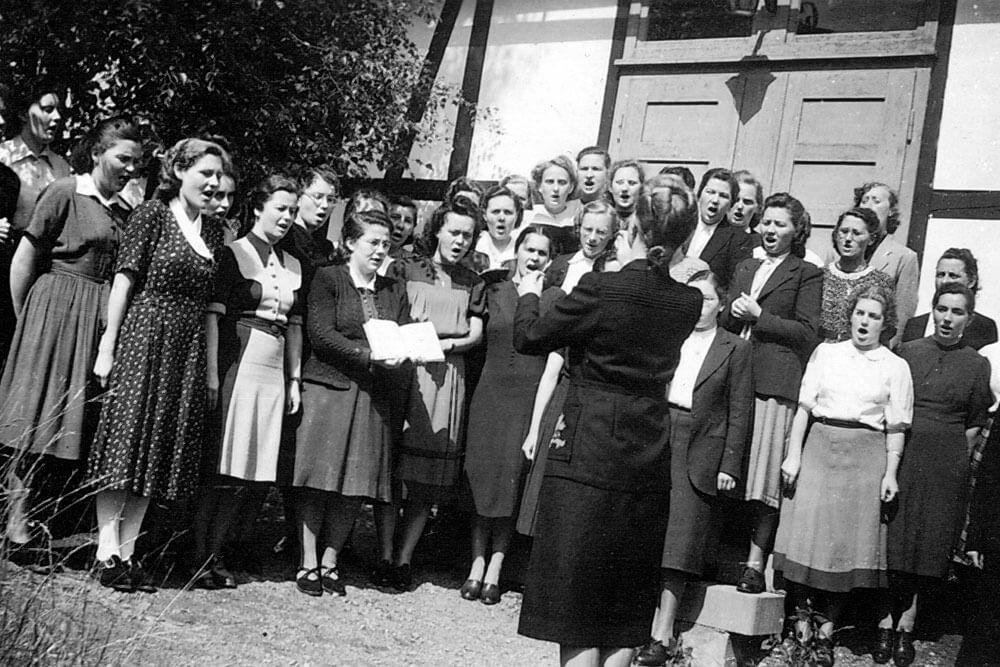 Historische Schwarz-Weiß-Fotografie eines Frauenchors bei einem Auftritt vor einem Fachwerkgebäude.