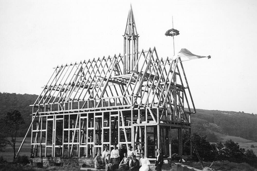 Historische Schwarz-Weiß-Fotografie eines Fachwerkbaus im Rohbau mit Richtkranz und mehreren Personen vor dem Gebäude.