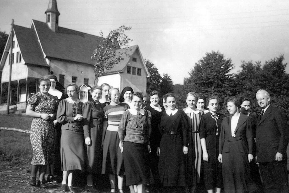 Historische Schwarz-Weiß-Fotografie einer Personengruppe vor einem Gebäude mit Kapelle in ländlicher Umgebung.