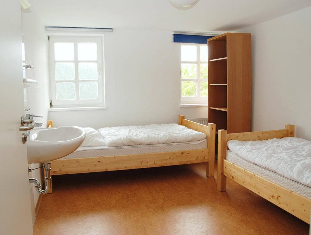 Helles Zweibettzimmer mit einfachen Holzmöbeln, Waschbecken, Regal und Blick ins Grüne.