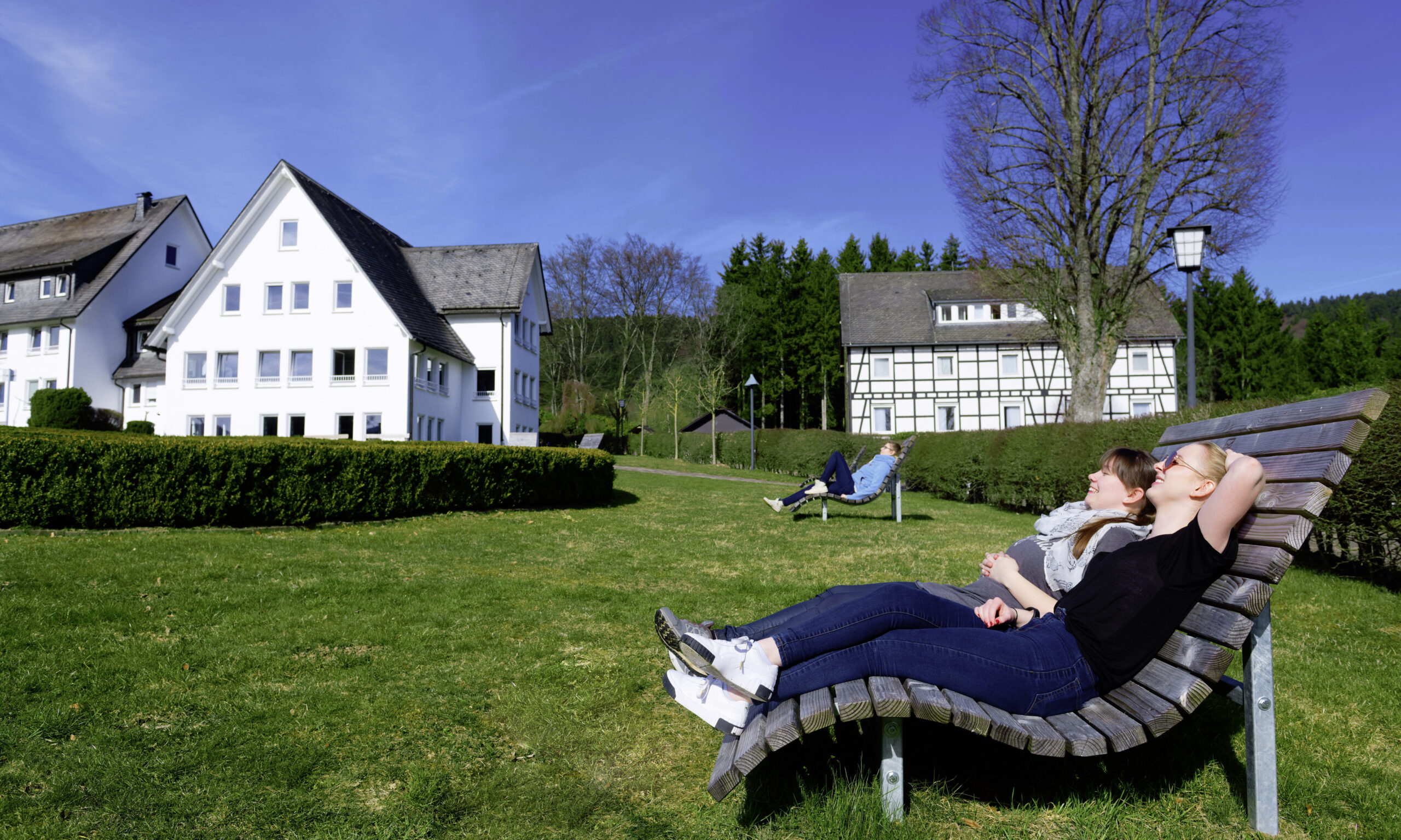 Zwei Frauen entspannen auf Holzliegen in einer gepflegten Grünanlage vor mehreren hellen Gebäuden; sie genießen die Sonne und blicken entspannt in den blauen Himmel, während im Hintergrund weitere Menschen und ein ruhiges, ländliches Umfeld mit Bäumen und Wiesen zu sehen sind.
