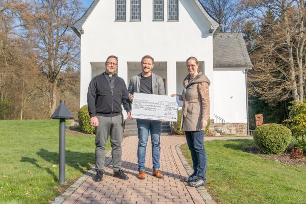 Foto vor der Kapelle mit großem Scheck. Dr. Andreas Rohde, Dominik Hansmeier (Caritasverband Meschede) und Anna Pietsch